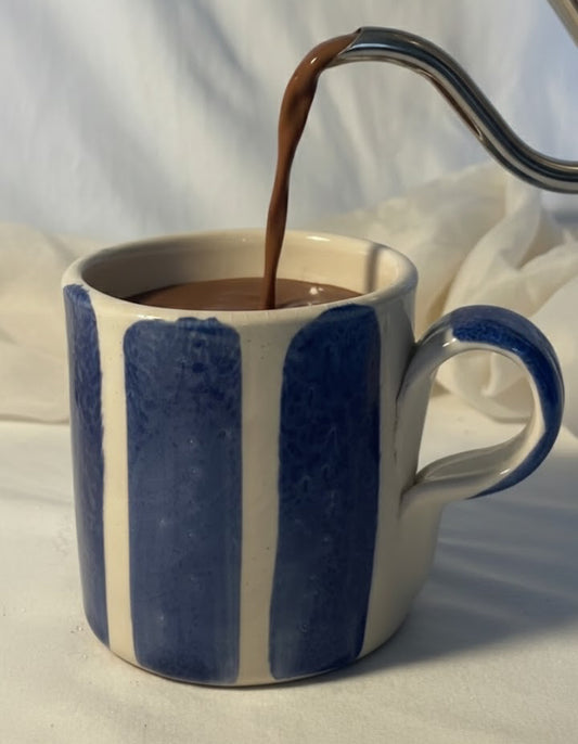 Heißer Kakao wird aus einer Silbernen Kanne in eine cremefarbene Tasse mit blauen Streifen gegossen, vor hellem Hintergrund.