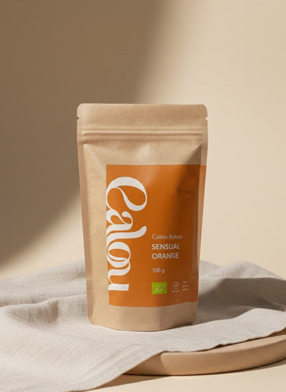 100 g Bio-Kakao ‚Calou Kakao Sensual Orange‘ in brauner Kraftpapierverpackung mit orangefarbenem Etikett, fotografiert auf beigem Tuch vor hellem, minimalistischen Hintergrund.