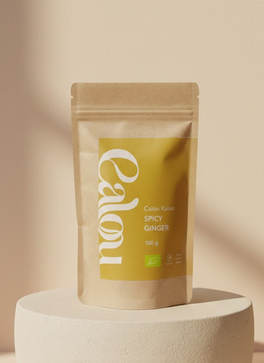 00 g Packung ‚Calou Kakao Spicy Ginger‘ in brauner Kraftpapierverpackung mit gelbem Etikett, stehend auf einem runden, cremefarbenen Podest vor hellem, minimalistischen Hintergrund.