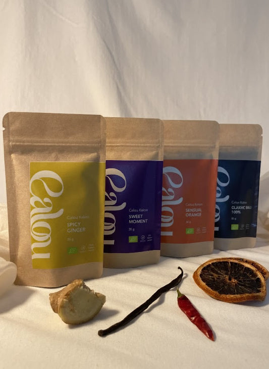 Vier Packungen Calou Kakao in verschiedenen Sorten stehen nebeneinander auf beigem Stoffhintergrund; davor liegen Ingwer, Vanilleschote, Chilischote und eine getrocknete Orangenscheibe. Warmes, weiches Studiolicht.